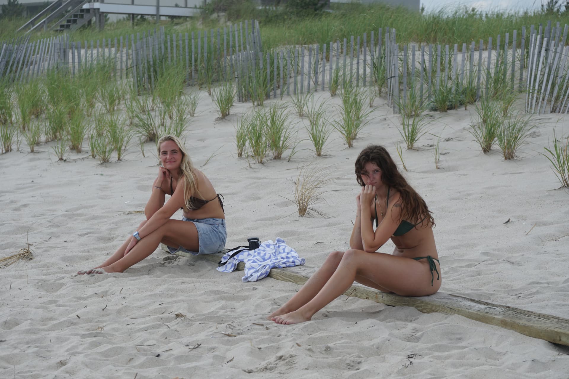 Two friends in bikinis sitting together on a weathered board in the sand, a towel between them